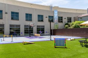 An outdoor recreational area features a basketball court, a Connect Four game, and cornhole boards on artificial grass in front of a building.
